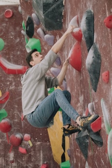 A person is engaged in indoor rock climbing on a wall covered with colorful climbing holds. The climber is wearing a gray hoodie, blue jeans, and climbing shoes, appearing focused and determined.