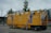 A large, mobile restroom unit with bright yellow walls and blue graphics. The unit is designed to serve both men and women, with doors labeled 'Damen' and 'Herren'. There are illustrations of a cartoon person in a uniform on the sides. The restroom is parked on a paved area with trees and a modern building in the background. Red and white caution tape and safety barriers are placed around it.