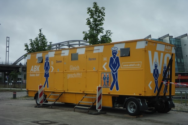 A large, mobile restroom unit with bright yellow walls and blue graphics. The unit is designed to serve both men and women, with doors labeled 'Damen' and 'Herren'. There are illustrations of a cartoon person in a uniform on the sides. The restroom is parked on a paved area with trees and a modern building in the background. Red and white caution tape and safety barriers are placed around it.