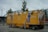 A large, mobile restroom unit with bright yellow walls and blue graphics. The unit is designed to serve both men and women, with doors labeled 'Damen' and 'Herren'. There are illustrations of a cartoon person in a uniform on the sides. The restroom is parked on a paved area with trees and a modern building in the background. Red and white caution tape and safety barriers are placed around it.