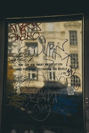 A glass surface covered in various colorful graffiti and writings with a reflection of a building in the background. The phrases and tags are layered, creating an urban and artistic look. The text partly reads 'YOU SAY YOU WANT MODERATION AND YOU WANT TO CHANGE THE WORLD.'