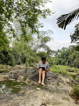 A person is sitting on a large rock in a lush, green natural environment, focusing on writing or drawing in a blue notebook. Surrounding them are trees with dense foliage and a small pool of water on the rock's surface, reflecting the greenery.