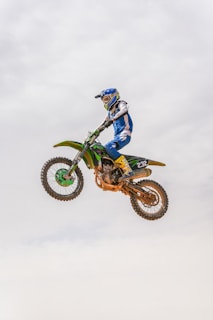 A person wearing blue and white motocross gear is performing a high jump on a green and black dirt bike. The bike is airborne against a clear sky, and the rider appears to be focused and in control.