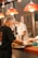 Cooks in a professional kitchen prepare and serve food under bright red pendant lights. They wear hairnets and face masks, indicating a focus on hygiene and safety. Steamers and other kitchen utensils are visible, suggesting a busy and efficient culinary environment.