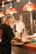 Cooks in a professional kitchen prepare and serve food under bright red pendant lights. They wear hairnets and face masks, indicating a focus on hygiene and safety. Steamers and other kitchen utensils are visible, suggesting a busy and efficient culinary environment.