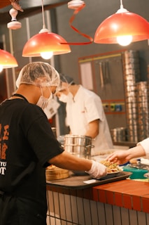 Cooks in a professional kitchen prepare and serve food under bright red pendant lights. They wear hairnets and face masks, indicating a focus on hygiene and safety. Steamers and other kitchen utensils are visible, suggesting a busy and efficient culinary environment.
