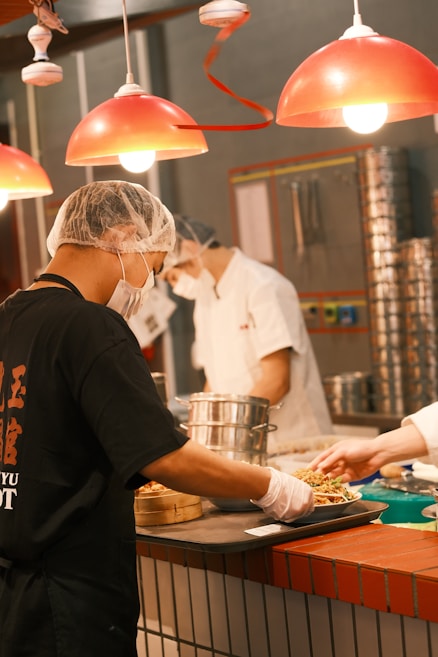 Cooks in a professional kitchen prepare and serve food under bright red pendant lights. They wear hairnets and face masks, indicating a focus on hygiene and safety. Steamers and other kitchen utensils are visible, suggesting a busy and efficient culinary environment.
