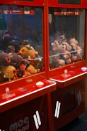 A pair of claw machines filled with colorful stuffed toys, featuring a variety of plush animals and characters. The machines are predominantly red, with clear glass panels and buttons on the front. The stuffed toys inside include a mix of colors, with some toys having features like large eyes and bright patterns.
