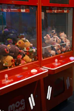 A pair of claw machines filled with colorful stuffed toys, featuring a variety of plush animals and characters. The machines are predominantly red, with clear glass panels and buttons on the front. The stuffed toys inside include a mix of colors, with some toys having features like large eyes and bright patterns.