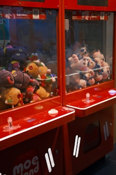 A pair of claw machines filled with colorful stuffed toys, featuring a variety of plush animals and characters. The machines are predominantly red, with clear glass panels and buttons on the front. The stuffed toys inside include a mix of colors, with some toys having features like large eyes and bright patterns.