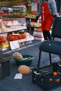 A street market scene with a variety of fruits displayed on shelves and tables. There are bananas and other fruits neatly arranged in the background. In the foreground, pineapples are placed in a basket on the ground, alongside a crate containing oranges. A person wearing a red apron and a checkered shirt stands nearby.