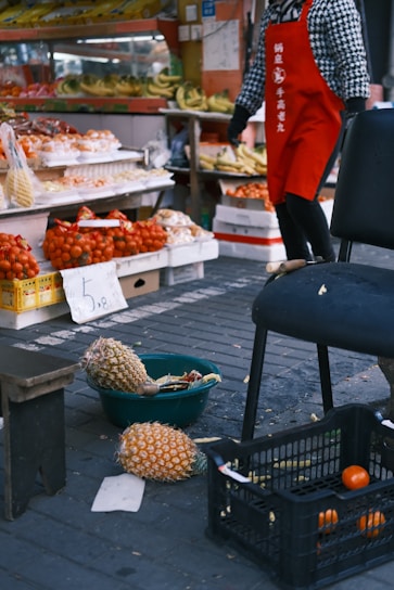 A street market scene with a variety of fruits displayed on shelves and tables. There are bananas and other fruits neatly arranged in the background. In the foreground, pineapples are placed in a basket on the ground, alongside a crate containing oranges. A person wearing a red apron and a checkered shirt stands nearby.