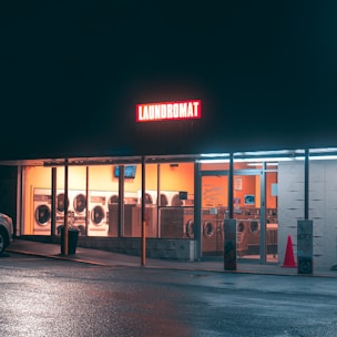 A brightly lit laundromat at night with a glowing red sign reading 'LAUNDROMAT'. The inside of the laundromat is illuminated with warm lighting, revealing rows of washing machines and dryers lining the walls. A cone and a trash can are visible outside the entrance, and the wet pavement reflects the lights from the building.
