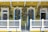A vibrant yellow house with white and blue trim features a charming porch adorned with an array of colorful floral wreaths hanging on each of the three doors. The decorative arrangement includes a variety of flowers and greenery, enhancing the cheerful and welcoming appearance of the house.