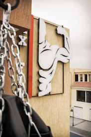 A stylized image of a flexing arm symbol is mounted on a beige building wall. In the foreground, chains hang down next to what appears to be a punching bag. The signage consists of bold black and white shapes, with a red vertical line accentuating the design. The setting is an urban environment with a nearby street and buildings visible in the background.