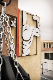 A stylized image of a flexing arm symbol is mounted on a beige building wall. In the foreground, chains hang down next to what appears to be a punching bag. The signage consists of bold black and white shapes, with a red vertical line accentuating the design. The setting is an urban environment with a nearby street and buildings visible in the background.