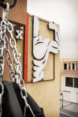 A stylized image of a flexing arm symbol is mounted on a beige building wall. In the foreground, chains hang down next to what appears to be a punching bag. The signage consists of bold black and white shapes, with a red vertical line accentuating the design. The setting is an urban environment with a nearby street and buildings visible in the background.