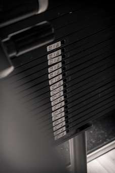 Multiple weight plates are stacked vertically, each labeled with weight in pounds and kilograms. The setup is likely part of a weightlifting or gym equipment, with a metal bar visible on the side.