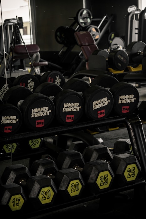 A gym setting featuring a variety of dumbbells neatly organized on a rack. The weights are labeled with different kilograms, including 25 and 20. In the background, there are larger gym machines and equipment, visible through dim lighting. The overall atmosphere suggests a space dedicated to fitness and strength training.