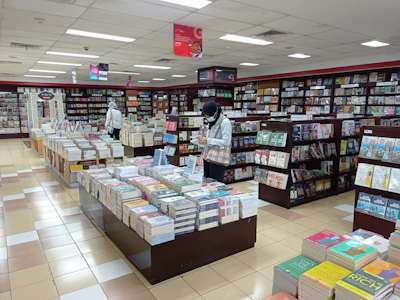 A bookstore filled with shelves and tables of neatly arranged books. Two people are browsing through the selection, one near a shelf and the other closer to the center table. The atmosphere is calm and organized, with bright overhead lighting illuminating the space.
