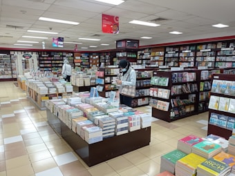 A bookstore filled with shelves and tables of neatly arranged books. Two people are browsing through the selection, one near a shelf and the other closer to the center table. The atmosphere is calm and organized, with bright overhead lighting illuminating the space.