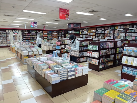 A bookstore filled with shelves and tables of neatly arranged books. Two people are browsing through the selection, one near a shelf and the other closer to the center table. The atmosphere is calm and organized, with bright overhead lighting illuminating the space.