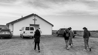 A black and white image depicting a group of people walking towards a small building with a cross on it. The building seems to be a church, and several vehicles, including a van and a pickup truck, are parked nearby. The scene is set in a rural area with a cloudy sky overhead.