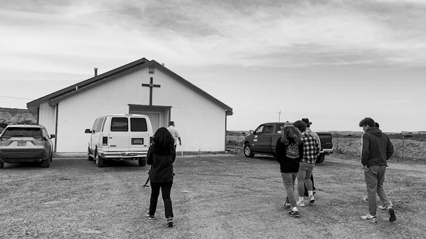 A black and white image depicting a group of people walking towards a small building with a cross on it. The building seems to be a church, and several vehicles, including a van and a pickup truck, are parked nearby. The scene is set in a rural area with a cloudy sky overhead.