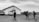A black and white image depicting a group of people walking towards a small building with a cross on it. The building seems to be a church, and several vehicles, including a van and a pickup truck, are parked nearby. The scene is set in a rural area with a cloudy sky overhead.