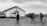 A black and white image depicting a group of people walking towards a small building with a cross on it. The building seems to be a church, and several vehicles, including a van and a pickup truck, are parked nearby. The scene is set in a rural area with a cloudy sky overhead.
