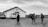 A black and white image depicting a group of people walking towards a small building with a cross on it. The building seems to be a church, and several vehicles, including a van and a pickup truck, are parked nearby. The scene is set in a rural area with a cloudy sky overhead.