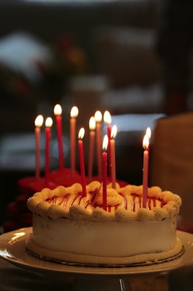 A round cake with white frosting and several lit red candles arranged on top, sitting on a white plate. The background is blurred, highlighting the cake as the focal point.