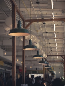 A busy indoor market or food court with people walking around and interacting. Large industrial-style green pendant lights hang from the ceiling, illuminating the area. The ceiling is filled with exposed pipes and ducts, adding to the industrial ambiance. Wooden beams separate different sections of the space.