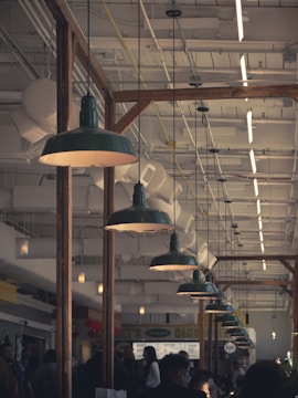 A busy indoor market or food court with people walking around and interacting. Large industrial-style green pendant lights hang from the ceiling, illuminating the area. The ceiling is filled with exposed pipes and ducts, adding to the industrial ambiance. Wooden beams separate different sections of the space.