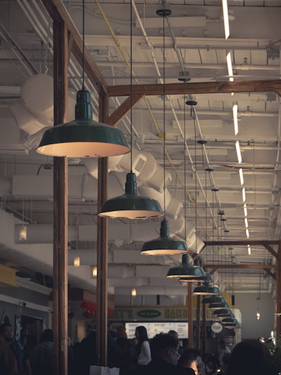 A busy indoor market or food court with people walking around and interacting. Large industrial-style green pendant lights hang from the ceiling, illuminating the area. The ceiling is filled with exposed pipes and ducts, adding to the industrial ambiance. Wooden beams separate different sections of the space.