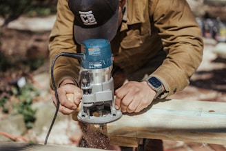 A person is using a handheld power tool on a wooden plank, focusing intently on the task. The individual is wearing a brown jacket, a cap, and a smartwatch, and is surrounded by an outdoor setting with blurred background elements.