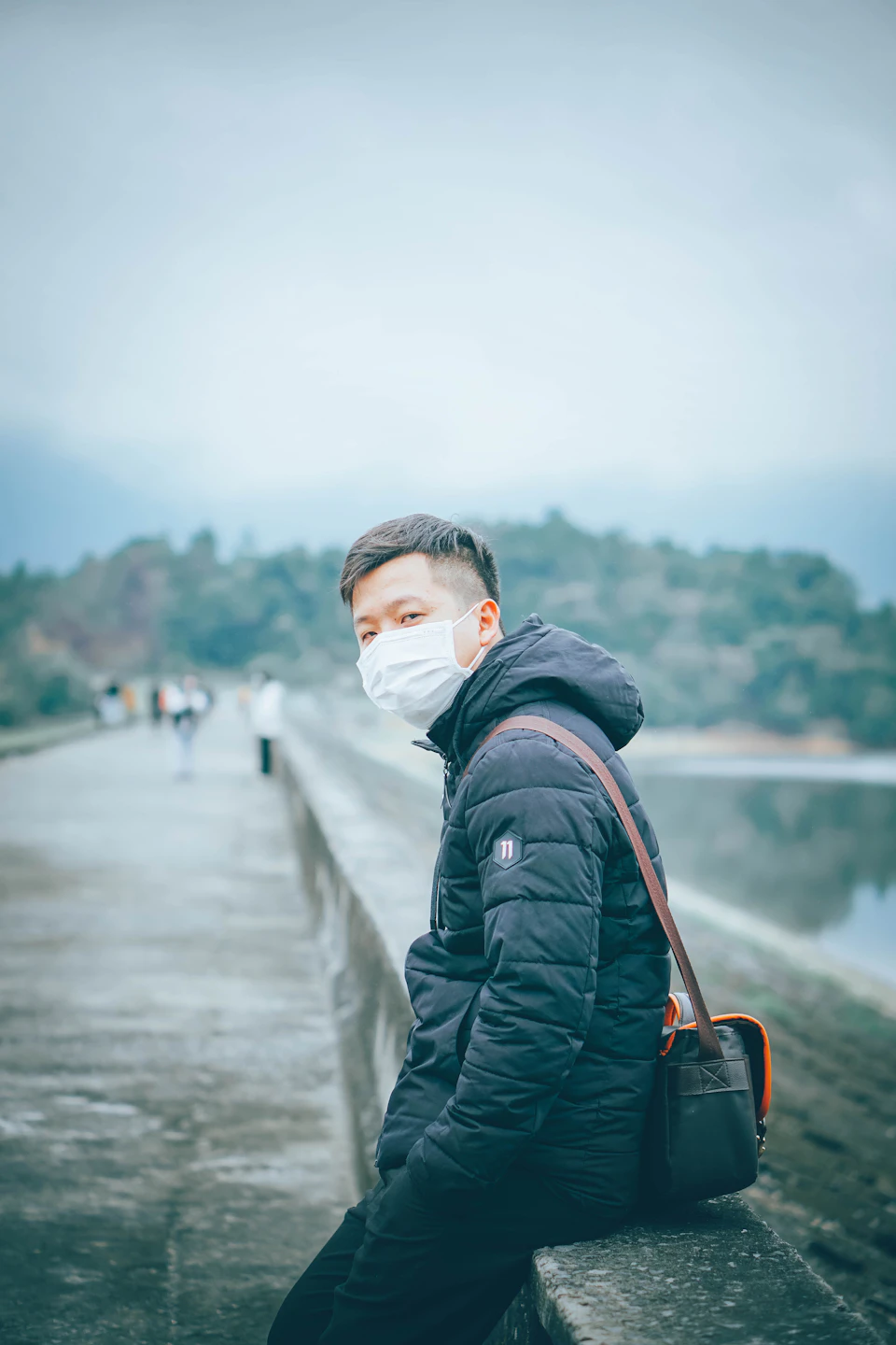 In the image, a young man is seen sitting on a concrete ledge or wall by a body of water. He's wearing a black jacket and a white face mask, which adds an air of mystery to his appearance. The man has a backpack slung over one shoulder, suggesting he might be out for a walk or hike. In the background, there are trees visible, indicating that they're in a natural setting. There are also several other people present in the scene, but their faces are blurred and indistinct.