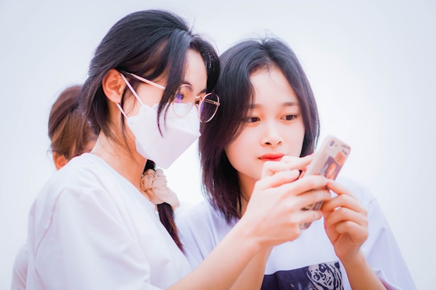 Two young women are looking at a smartphone together. One of them is wearing glasses and a face mask, while both are wearing casual white clothing. Both appear engaged and focused on the phone screen as they browse something together.