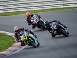 Three motorcyclists are racing around a track, leaning sharply into a curve. The riders wear helmets and racing suits, displaying focus and precision as they navigate the turn. The motorcycles are colorful and branded, emphasizing speed and competition.
