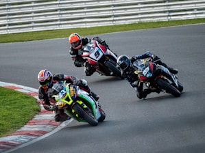 Three motorcyclists are racing around a track, leaning sharply into a curve. The riders wear helmets and racing suits, displaying focus and precision as they navigate the turn. The motorcycles are colorful and branded, emphasizing speed and competition.