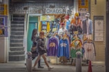 A street-side clothing shop displaying colorful sportswear and jackets on mannequins. Two mannequins are fully dressed with caps, while the others display clothing items on hangers. The shop is named 'CHAOS' and has bright signs with text and graphics. Two women in winter attire walk past the store, and a staircase is visible on the left.