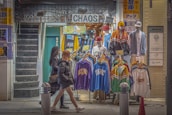 A street-side clothing shop displaying colorful sportswear and jackets on mannequins. Two mannequins are fully dressed with caps, while the others display clothing items on hangers. The shop is named 'CHAOS' and has bright signs with text and graphics. Two women in winter attire walk past the store, and a staircase is visible on the left.
