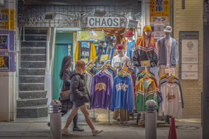 A street-side clothing shop displaying colorful sportswear and jackets on mannequins. Two mannequins are fully dressed with caps, while the others display clothing items on hangers. The shop is named 'CHAOS' and has bright signs with text and graphics. Two women in winter attire walk past the store, and a staircase is visible on the left.