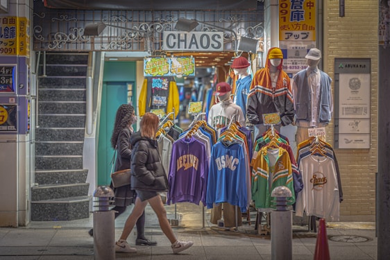 A street-side clothing shop displaying colorful sportswear and jackets on mannequins. Two mannequins are fully dressed with caps, while the others display clothing items on hangers. The shop is named 'CHAOS' and has bright signs with text and graphics. Two women in winter attire walk past the store, and a staircase is visible on the left.