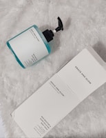 A bottle of body wash with a pump dispenser sits on a fluffy white surface. The bottle is translucent with blue liquid inside and features a label reading 'Grace and Glow.' Next to it lies its matching product packaging box, also labeled with the same brand name and product details.