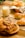 A selection of freshly baked pastries, including croissants and rolls, arranged on a wooden serving board and a wire rack. The pastries are golden-brown, hinting at their flaky texture, with some topped with sesame seeds or chocolate chips. A glass of milk is placed nearby, adding a touch of comfort and homeliness. The background includes a checkered cloth and a wooden surface, enhancing the cozy breakfast setting.