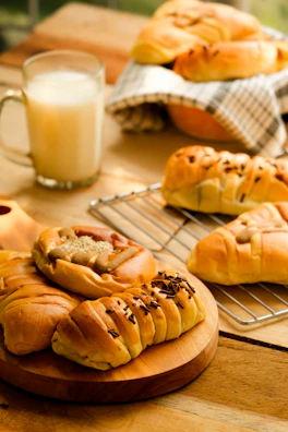 A selection of freshly baked pastries, including croissants and rolls, arranged on a wooden serving board and a wire rack. The pastries are golden-brown, hinting at their flaky texture, with some topped with sesame seeds or chocolate chips. A glass of milk is placed nearby, adding a touch of comfort and homeliness. The background includes a checkered cloth and a wooden surface, enhancing the cozy breakfast setting.
