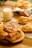 A selection of freshly baked pastries, including croissants and rolls, arranged on a wooden serving board and a wire rack. The pastries are golden-brown, hinting at their flaky texture, with some topped with sesame seeds or chocolate chips. A glass of milk is placed nearby, adding a touch of comfort and homeliness. The background includes a checkered cloth and a wooden surface, enhancing the cozy breakfast setting.