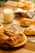 A selection of freshly baked pastries, including croissants and rolls, arranged on a wooden serving board and a wire rack. The pastries are golden-brown, hinting at their flaky texture, with some topped with sesame seeds or chocolate chips. A glass of milk is placed nearby, adding a touch of comfort and homeliness. The background includes a checkered cloth and a wooden surface, enhancing the cozy breakfast setting.