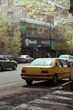 A yellow taxi is parked on the side of a city street, with partially visible license plates. Surrounding the taxi are other parked cars and a building with a shop named 'Benelli'. The trees have budding green leaves, indicating springtime.